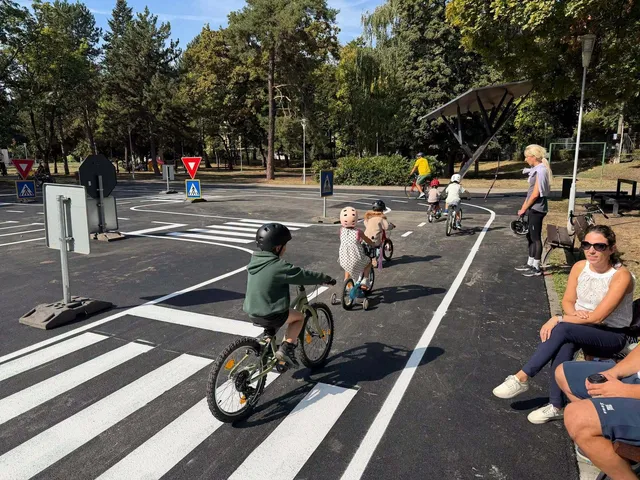 Copii pe biciclete circulă pe un poligon asfaltat marcat cu linii albe, treceri de pietoni și semne rutiere sub copaci.