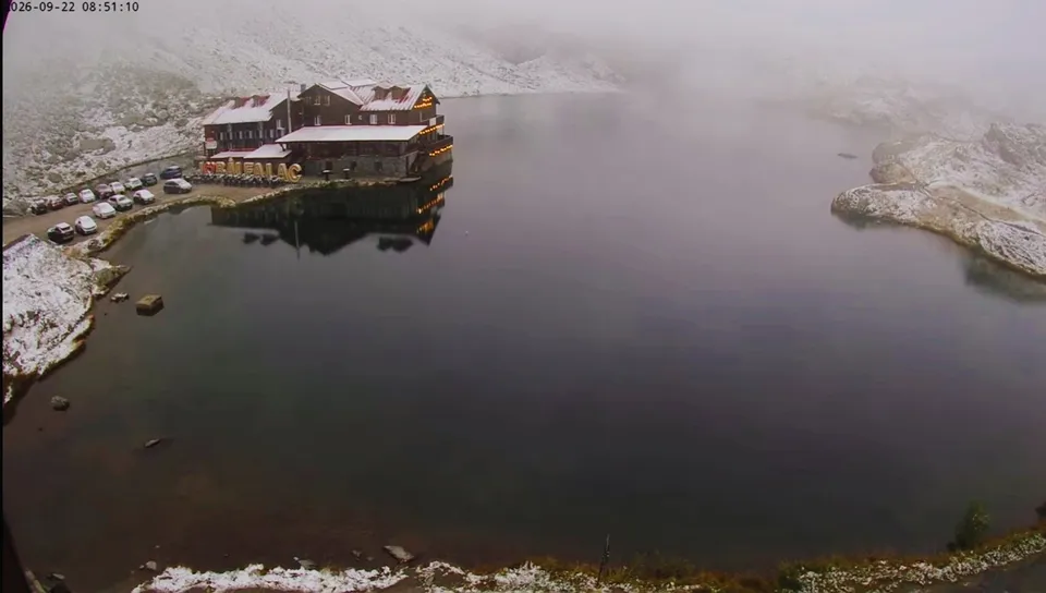 Un lac întins este înconjurat de munți acoperiți de zăpadă și de o clădire cu acoperiș înclinat lângă un parc de mașini.