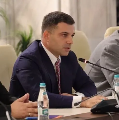 Un bărbat cu părul scurt, purtând un costum închis la culoare și o cravată modelată, stă la o masă lângă un microfon.