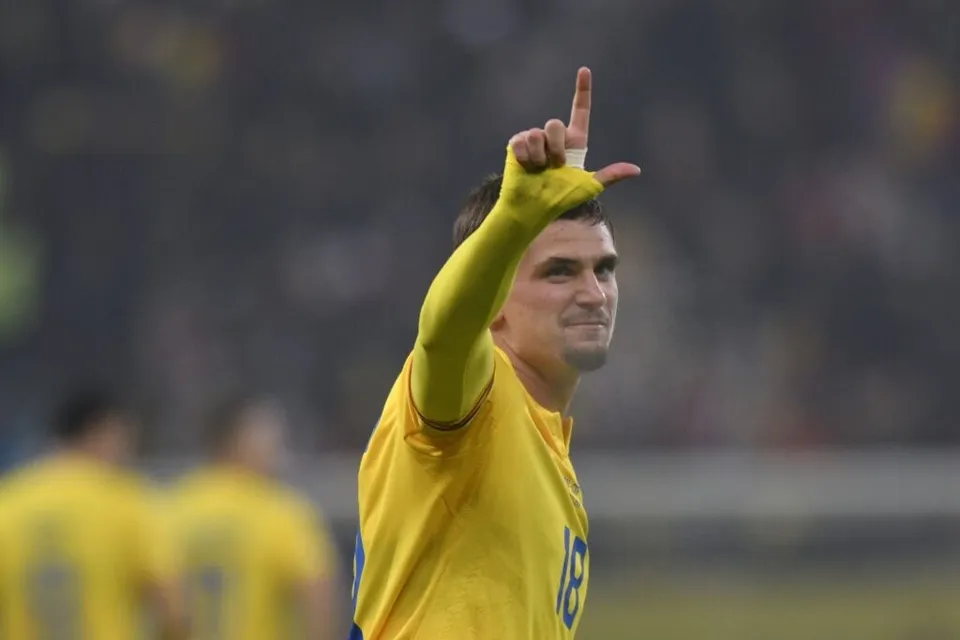 Un bărbat cu tricou galben ridică mâna dreaptă cu degetul arătător spre cer pe un teren de fotbal.