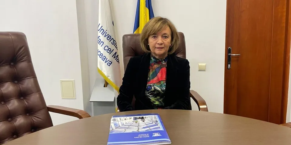 O femeie cu părul scurt și blond stă la o masă de lemn lângă un document albastru într-o cameră cu un steag în fundal.