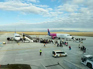 Avioane sunt parcate pe pista unui aeroport, în timp ce grupuri de oameni stau lângă scări mobile și vehicule de serviciu pe asfaltul deschis.