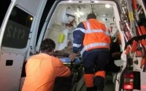 Două persoane purtând haine reflectorizante orange și albastre se află lângă spatele unei ambulanțe deschise.