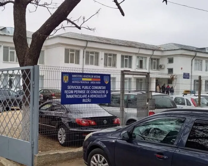 O clădire albă cu ferestre este situată în spatele unui gard metalic, având mașini parcate în fața acesteia.