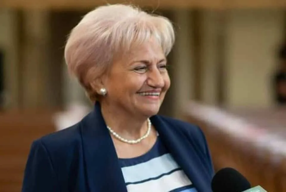 O femeie cu părul scurt și deschis la culoare poartă un lănț de perle și un sacou albastru în fața unui fundal neclar.