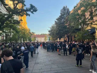 O mulțime de oameni stă în picioare pe o stradă pietonală cu clădiri și copaci pe ambele părți.