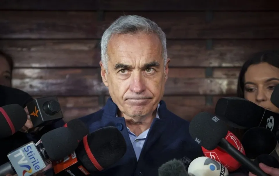 Un bărbat cu părul cărunt și o jachetă albastră este înconjurat de mai multe microfoane de la conferința de presă.
