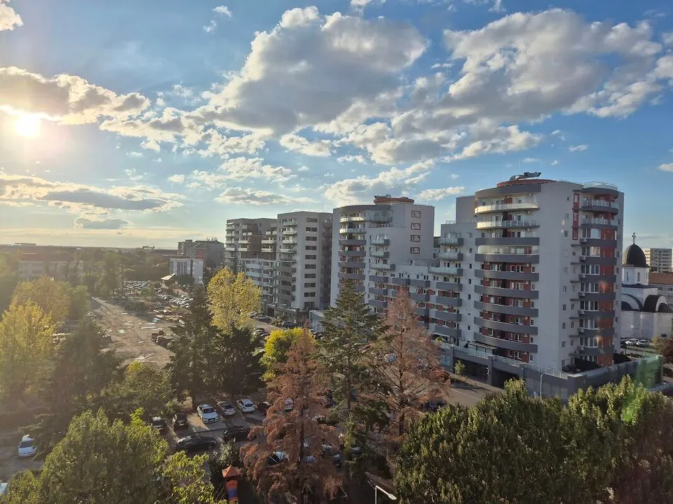 Blocuri înalte de apartamente sunt situate lângă copaci verzi și maronii sub un cer cu nori și soare.