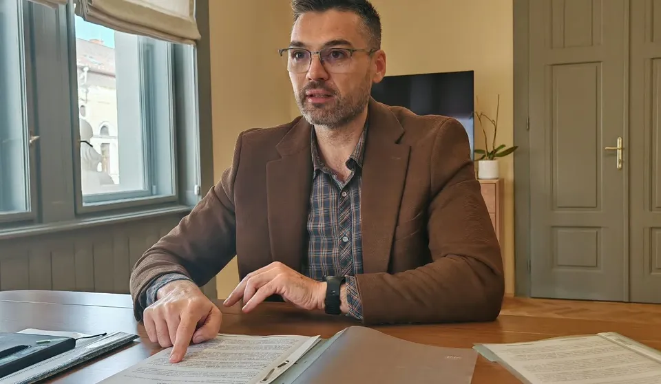 Un bărbat cu ochelari și sacou maro stă la o masă de lemn, arătând cu degetul spre un document de pe suprafață.