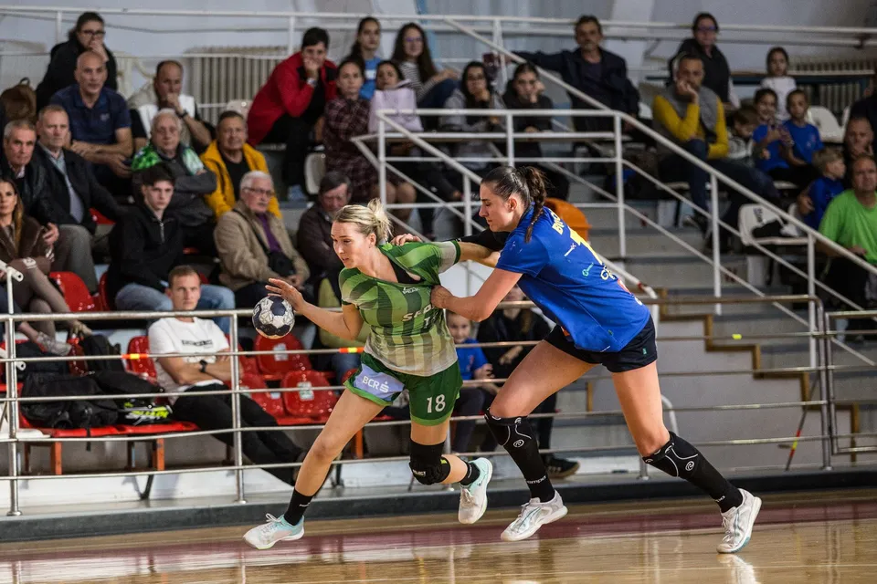 Două jucătoare de handbal se confruntă pe terenul unei săli de sport, în timp ce spectatori stau pe tribunele din fundal.