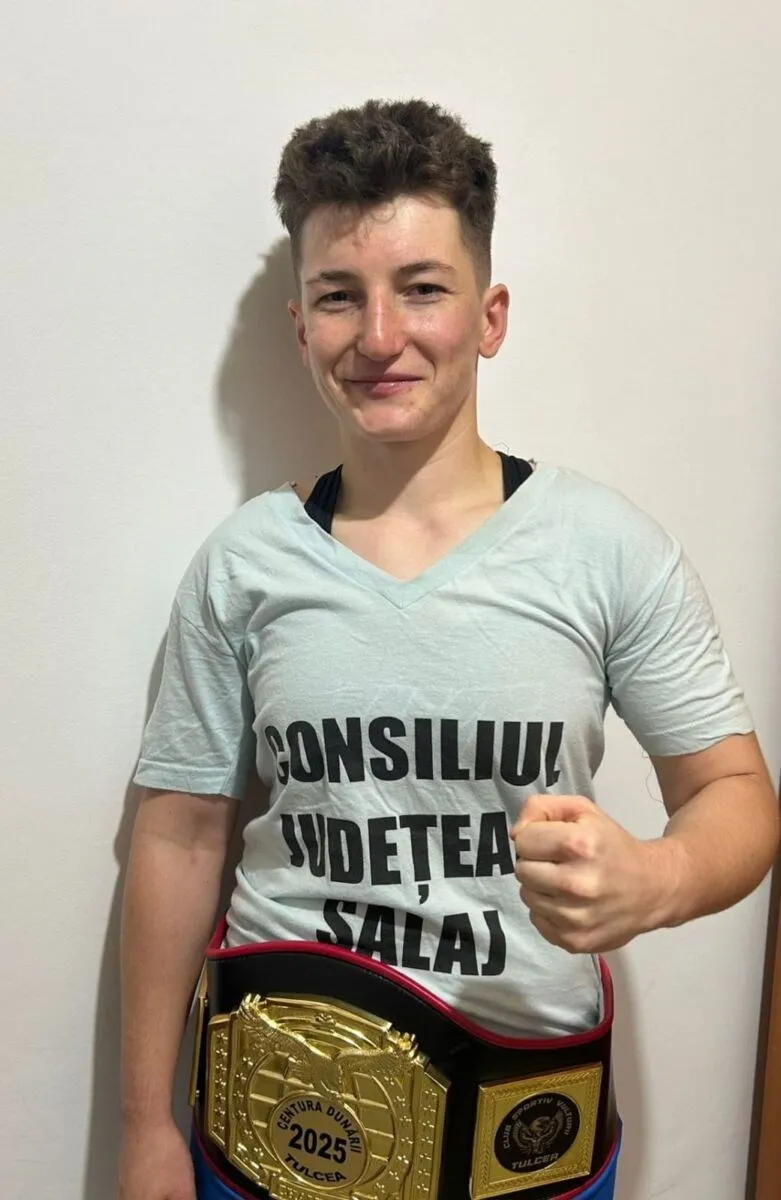 O persoană zâmbește purtând un tricou deschis la gât cu text negru și un centură de campion cu plăci aurii în partea inferioară.