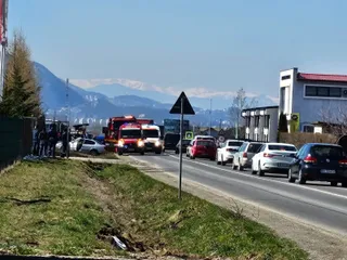 Vehicule de serviciu și mașini sunt parcate pe marginea unui drum asfaltat, sub un cer senin, în fața unor munți înzăpeziți.