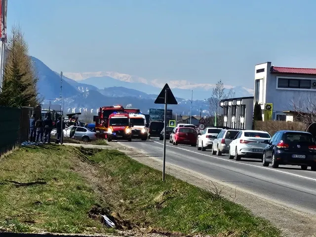 Vehicule de serviciu și mașini sunt parcate pe marginea unui drum asfaltat, sub un cer senin, în fața unor munți înzăpeziți.
