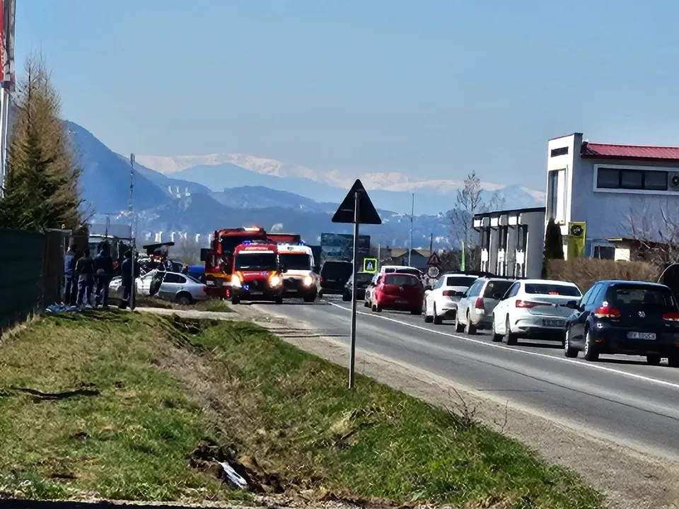 Vehicule de serviciu și mașini sunt parcate pe marginea unui drum asfaltat, sub un cer senin, în fața unor munți înzăpeziți.