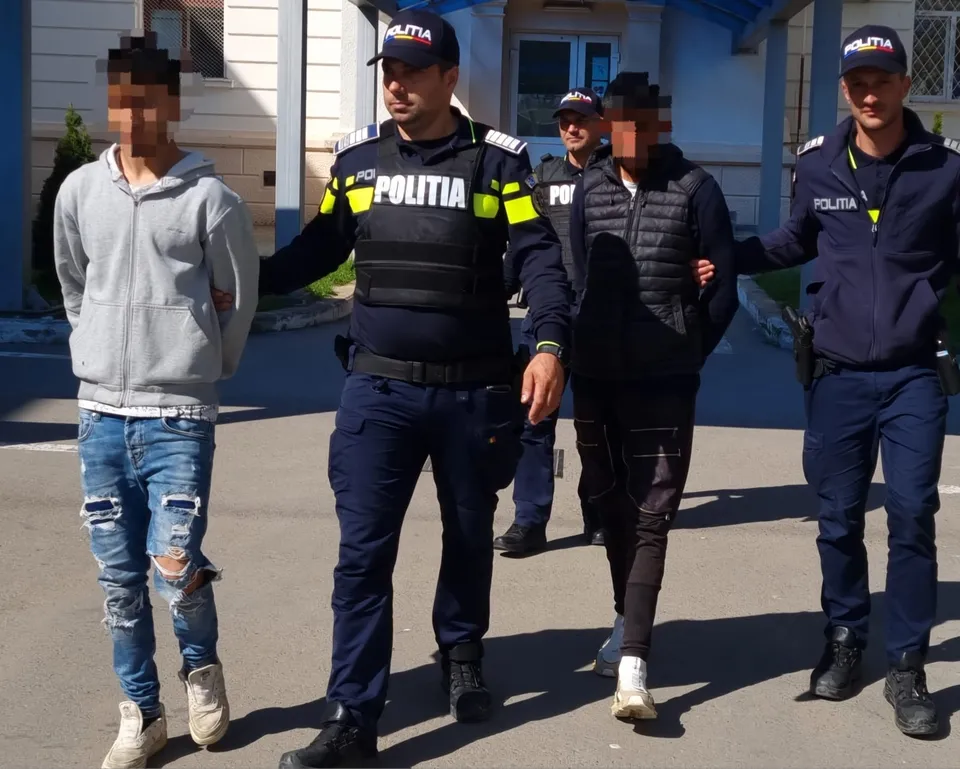 Patru bărbați merg pe o stradă, doi dintre ei purtând uniforme de polițist și conducând doi tineri cu fețele pixelate.