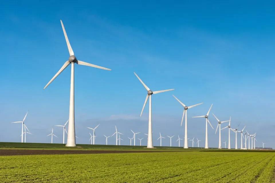Mai multe turbine eoliene albe sunt amplasate într-un câmp verde sub un cer albastru luminos.
