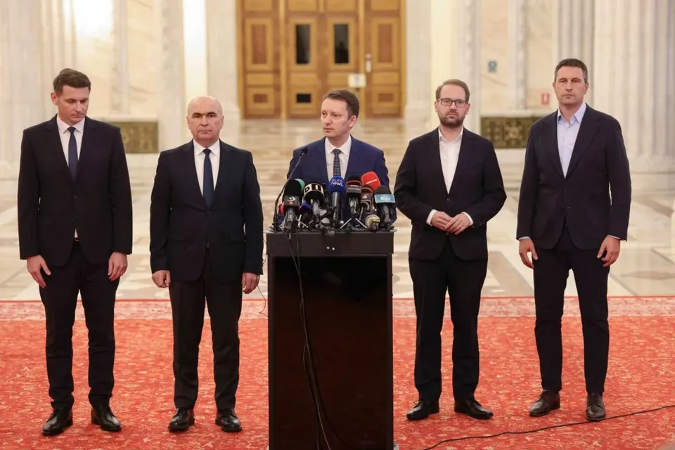 Cinci bărbați în costume scurte stau în picioare în spatele unui podium cu microfoane pe un covor roșu.