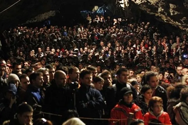 O mulțime de oameni stă concentrată într-un spațiu întunecat cu pereți de stâncă, privind spre o direcție comună.