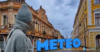 O persoană cu căciulă gri și haine gri privește spre o stradă cu clădiri galbene sub un cer înnorat.