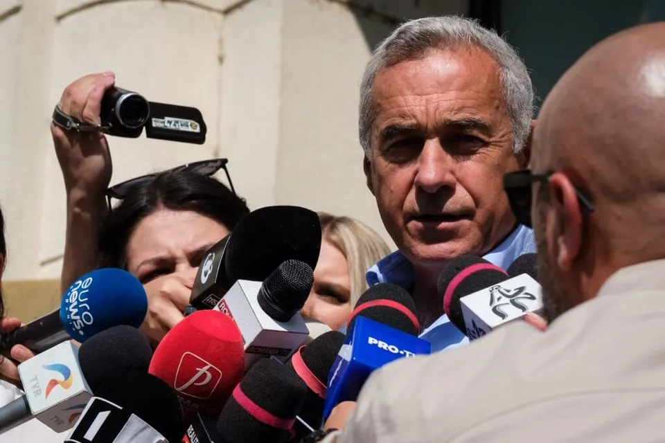 Un bărbat cu părul cărunt este înconjurat de microfoane și o cameră video în timp ce o persoană cu ochelari îi stă în față.