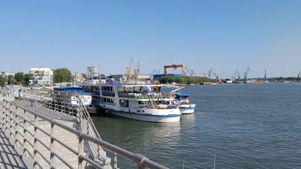 Mai multe nave albe sunt ancorate pe un fluviu lângă un mal cu un parapet metalic sub un cer senin.
