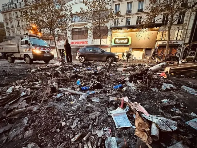 Un drum urban este acoperit de resturi, deșeuri și obiecte incendiate în fața unor magazine și a unor mașini parcate.