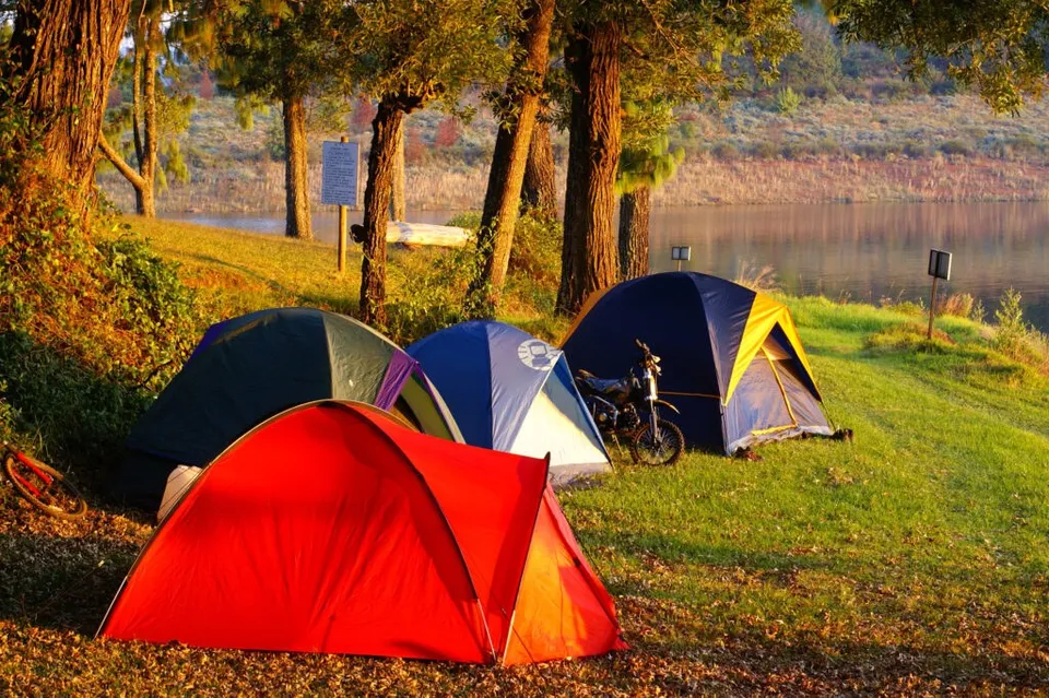 Trei corturi de camping de diferite culori sunt amplasate pe o iarbă verde lângă un lac, lângă un motocicletă parcată.