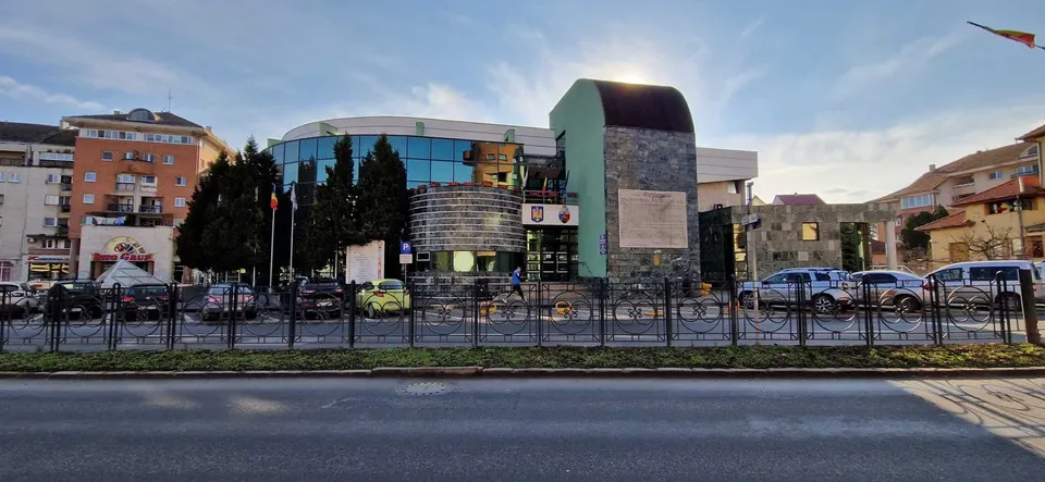 O clădire modernă cu fațadă de sticlă și un element vertical masiv din piatră se află în spatele unui gard metalic și a unor mașini parcate.