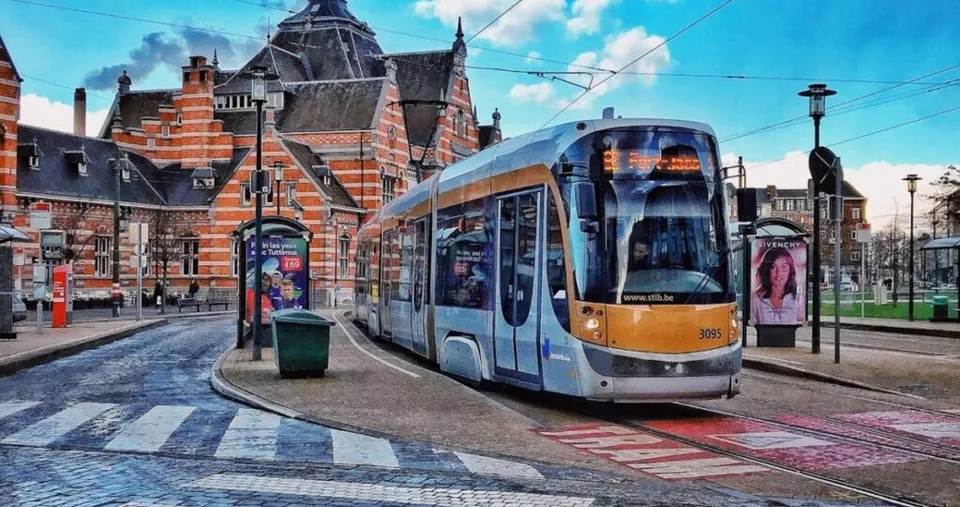 Un tramvai modern de culoare gri și galben circulă pe o cale ferată urbană lângă clădiri din cărămidă roșie sub un cer înnorat.