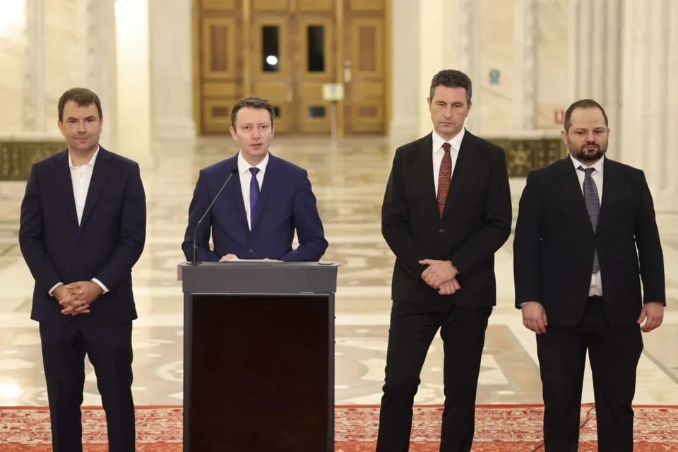 Patru bărbați în costume scurte stau în picioare într-o încăpere cu coloane și uși mari de lemn, unul fiind poziționat în spatele unui podium.