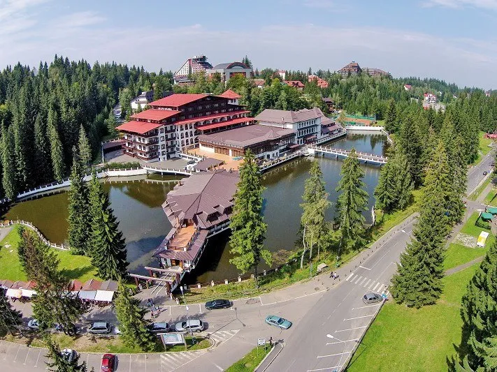 O vedere aeriană prezintă clădiri de hotel înconjurate de pădure și un lac artificial cu un pod peste acesta.