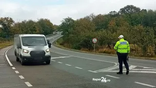 Un agent de poliție în uniformă reflectorizantă verde stă pe marginea unei drumuri asfaltate lângă un van alb care circulă pe banda de circulație.