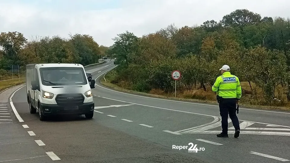 Un agent de poliție în uniformă reflectorizantă verde stă pe marginea unei drumuri asfaltate lângă un van alb care circulă pe banda de circulație.