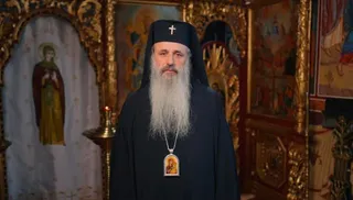 Un bărbat cu barbă lungă și albă poartă o mantie neagră, un hat negru cu o cruce și un medalion cu o icoană.