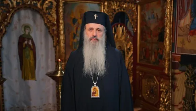 Un bărbat cu barbă lungă și albă poartă o mantie neagră, un hat negru cu o cruce și un medalion cu o icoană.