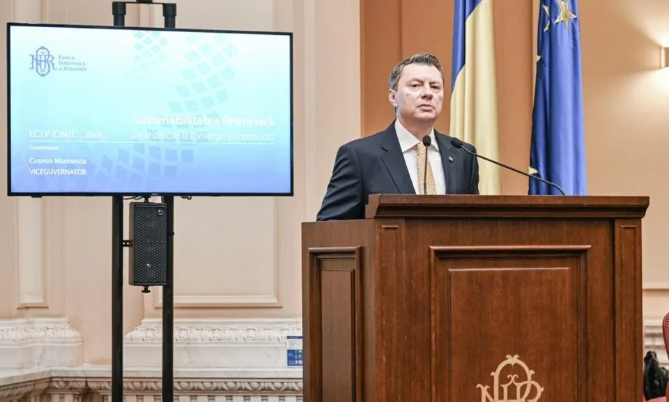 Un bărbat într-un costum închis la culoare stă în spatele unui podium de lemn lângă un ecran de monitor care afișează text și un logo.