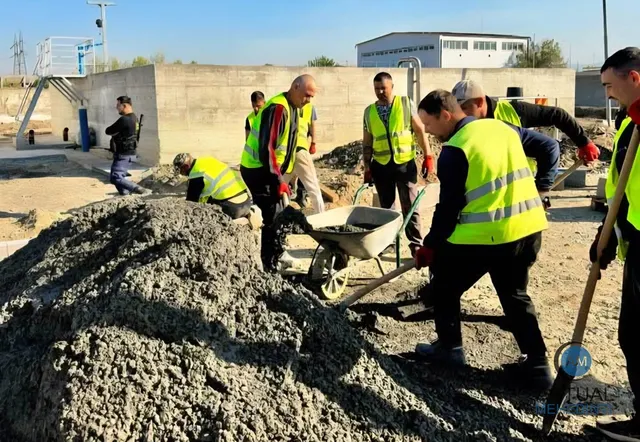 Mai mulți bărbați purtând veste de siguranță galbenă lucrează cu un cărucior și un morman de nisip într-un șantier aflat în aer liber.