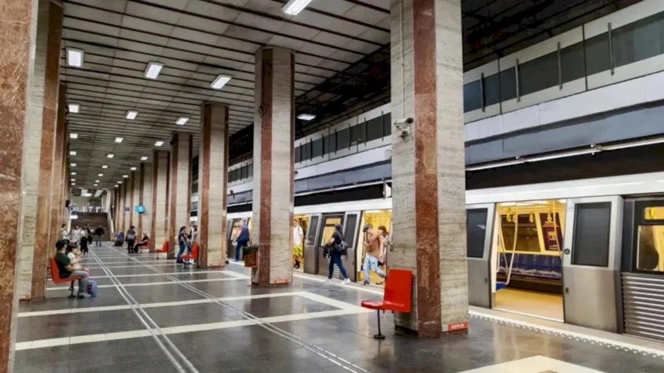 O platformă de metrou cu coloane rectangulare, scaune roșii și un tren cu ușile deschise pe care se văd pasageri.