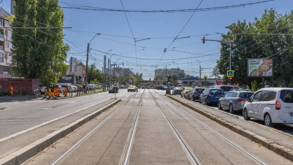 O stradă cu șine de tramvai centrale, flancată de mașini parcate și clădiri sub un cer senin.