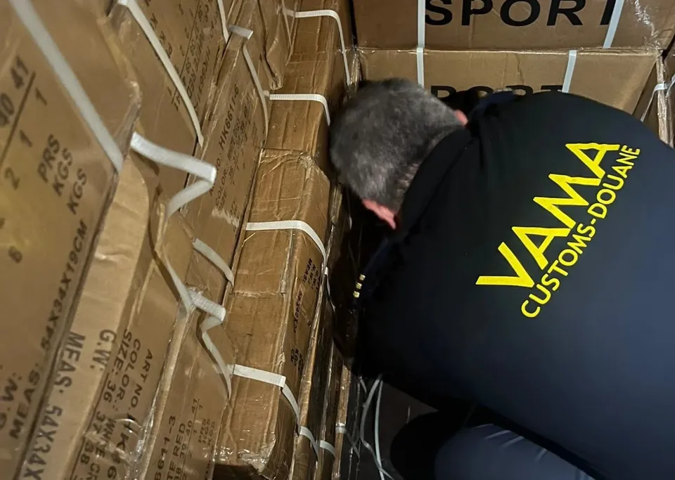 Un bărbat cu un tricou negru pe care scrie VAMA CUSTOMS-DOUANE este aplecat între cutii de carton maronii stivuite într-un vehicul.
