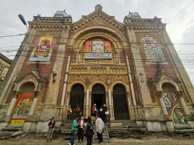 O clădire veche din cărămidă cu trei arcade centrale și decoruri sculptate prezintă diverse afișe colorate aplicate pe fațadă în dreptul ferestrelor și porților.
