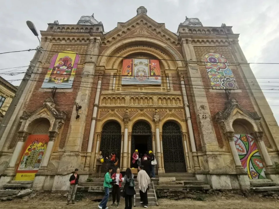 O clădire veche din cărămidă cu trei arcade centrale și decoruri sculptate prezintă diverse afișe colorate aplicate pe fațadă în dreptul ferestrelor și porților.