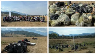 Un grup mare de oameni stă întrupat într-un câmp deschis, lângă saci de gunoi negri și saci de plastic plini, sub un cer senin.