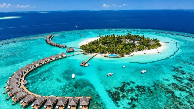O vedere aeriană a unei insule tropicale cu vegetație verde, plaje albe și un șir de cabane cu acoperiș de paie dispuse pe un ponton de lemn.