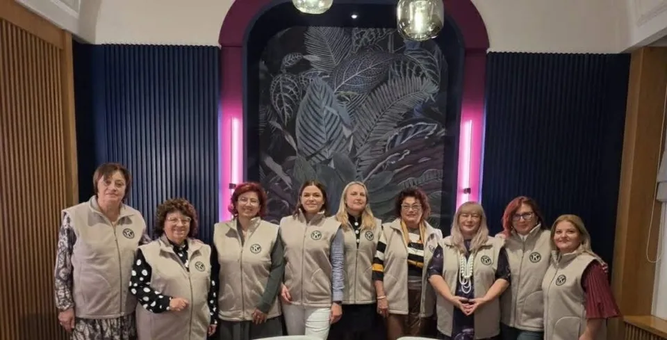 Nouă femei poartă veste de culoare deschisă și stau în rând în fața unui perete decorat cu motive vegetale și lumini roz.