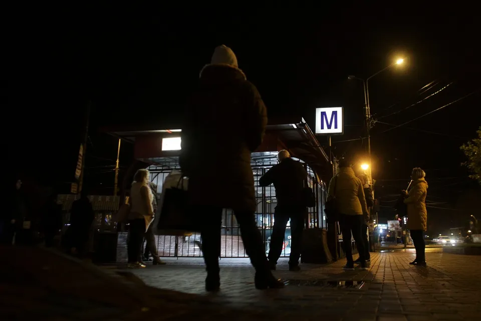 Siluete de persoane merg pe un trotuar noaptea lângă o intrare de metrou marcată cu un semn alb cu litera M albastră.