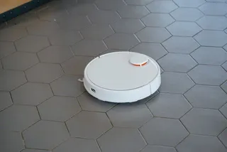 Un aspirator robot alb este poziționat pe un podea formată din plăci hexagonale gri.