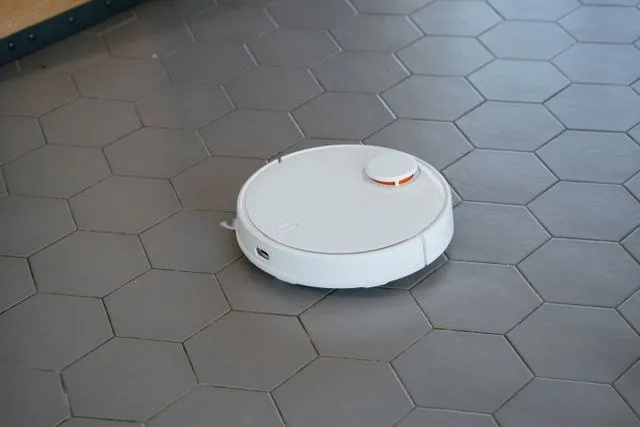 Un aspirator robot alb este poziționat pe un podea formată din plăci hexagonale gri.