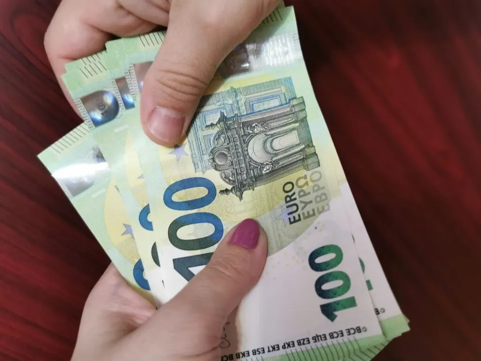 Două mâini țin un teanc de bancnote de o sută de euro pe o suprafață de culoare roșie.