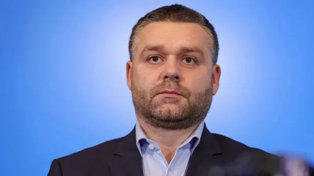 Un bărbat cu barbă scurtă și păr scurt poartă o cămașă deschisă la guler și un sacou în fața unui fundal albastru.